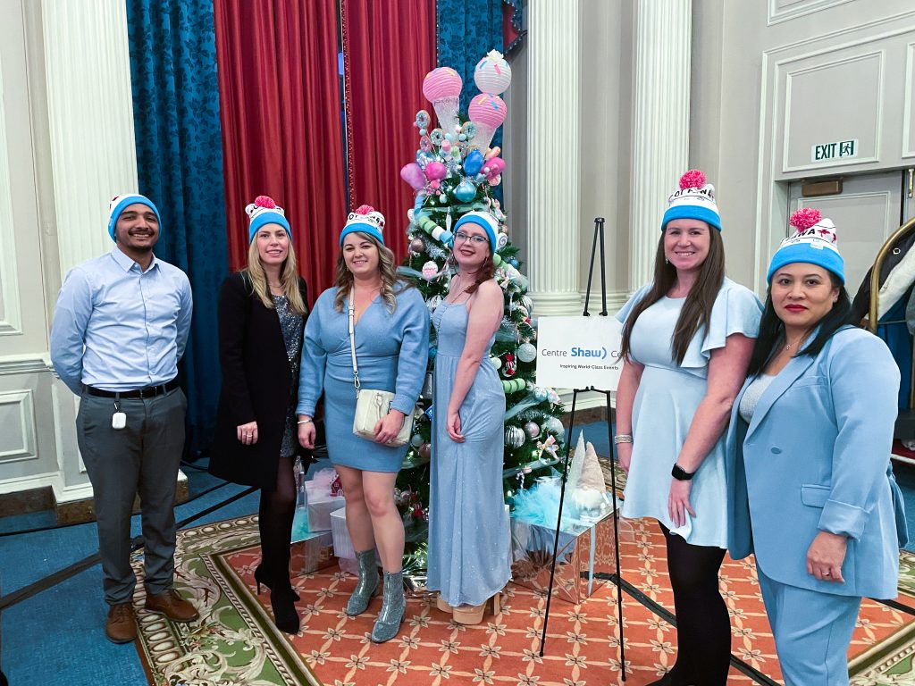 Shaw Centre team stands infront of Christmas tree in patching pastel blue outfits and Shaw Centre toques